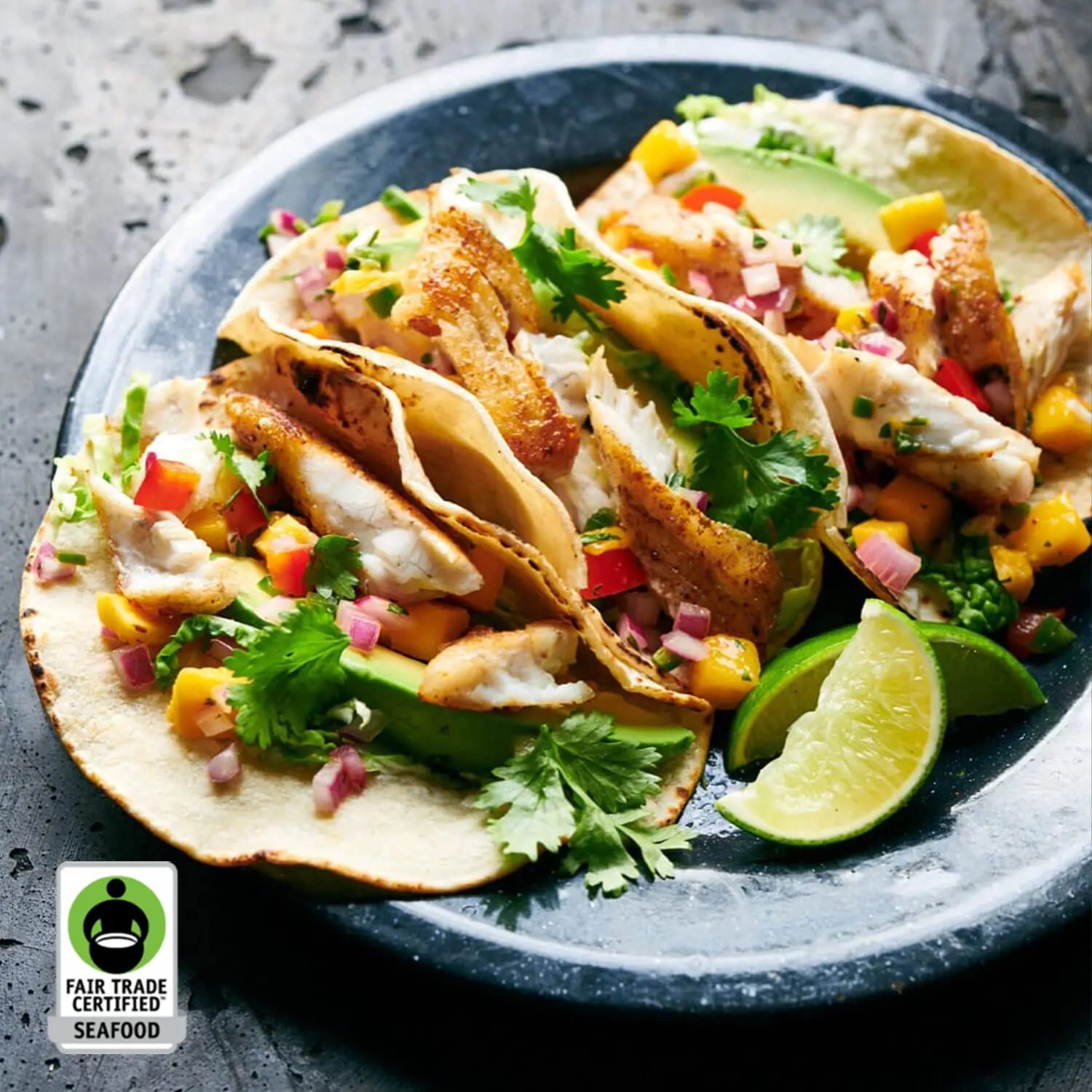 Four fish tacos filled with white fish, topped with diced red and yellow peppers, cilantro, and avocado on a dark plate, with lime wedges and a Fair Trade Certified Seafood badge in the corner.